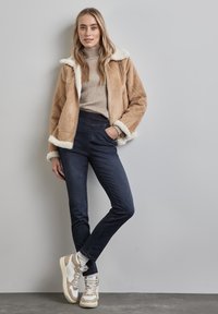 Tan faux shearlingjacka med vit foder, beige ribbad polotröja, mörkblå jeans och vita sneakers med beiga detaljer.