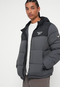 Doplnková zateplená bunda v tmavo šedej farbe s čiernou kapucňou, so zipsom, elastickými manžetami a logom Reebok na hrudi a rukáve.