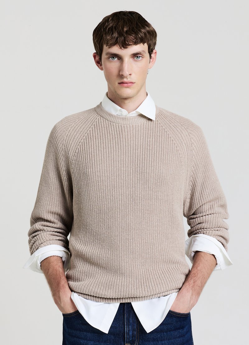 Jeune homme aux cheveux bruns courts portant un pull en tricot beige par-dessus une chemise blanche avec les manches retroussées, mains dans les poches, fond neutre.