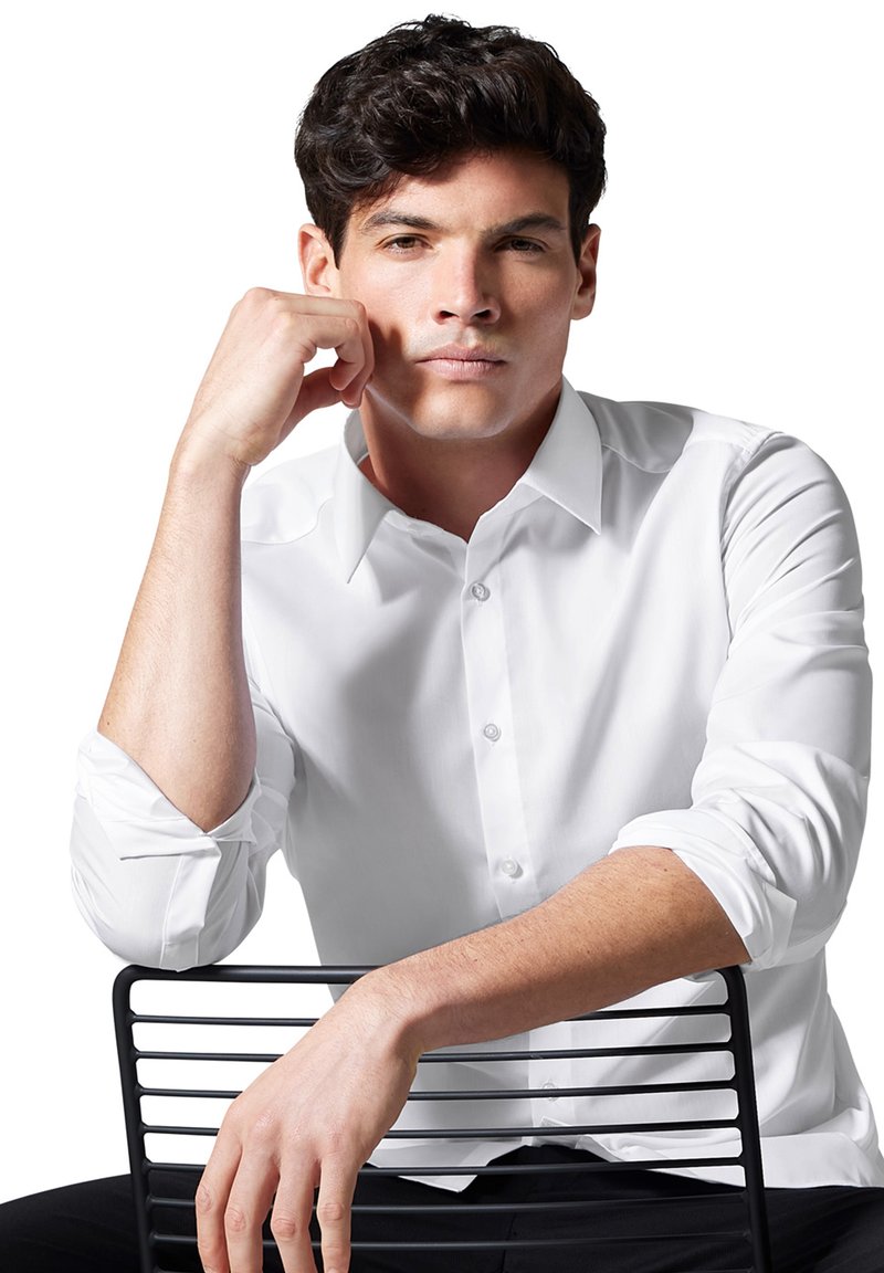 Weißes Langarmshirt mit Button-Down-Kragen, das eine glatte Textur aufweist. Modell sitzt auf einem schwarzen Metallstuhl und schlägt eine entspannte Pose ein.