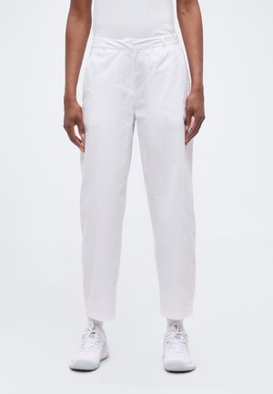 Personne portant un pantalon blanc ample, des chaussettes blanches et des baskets blanches, debout devant un fond uni.