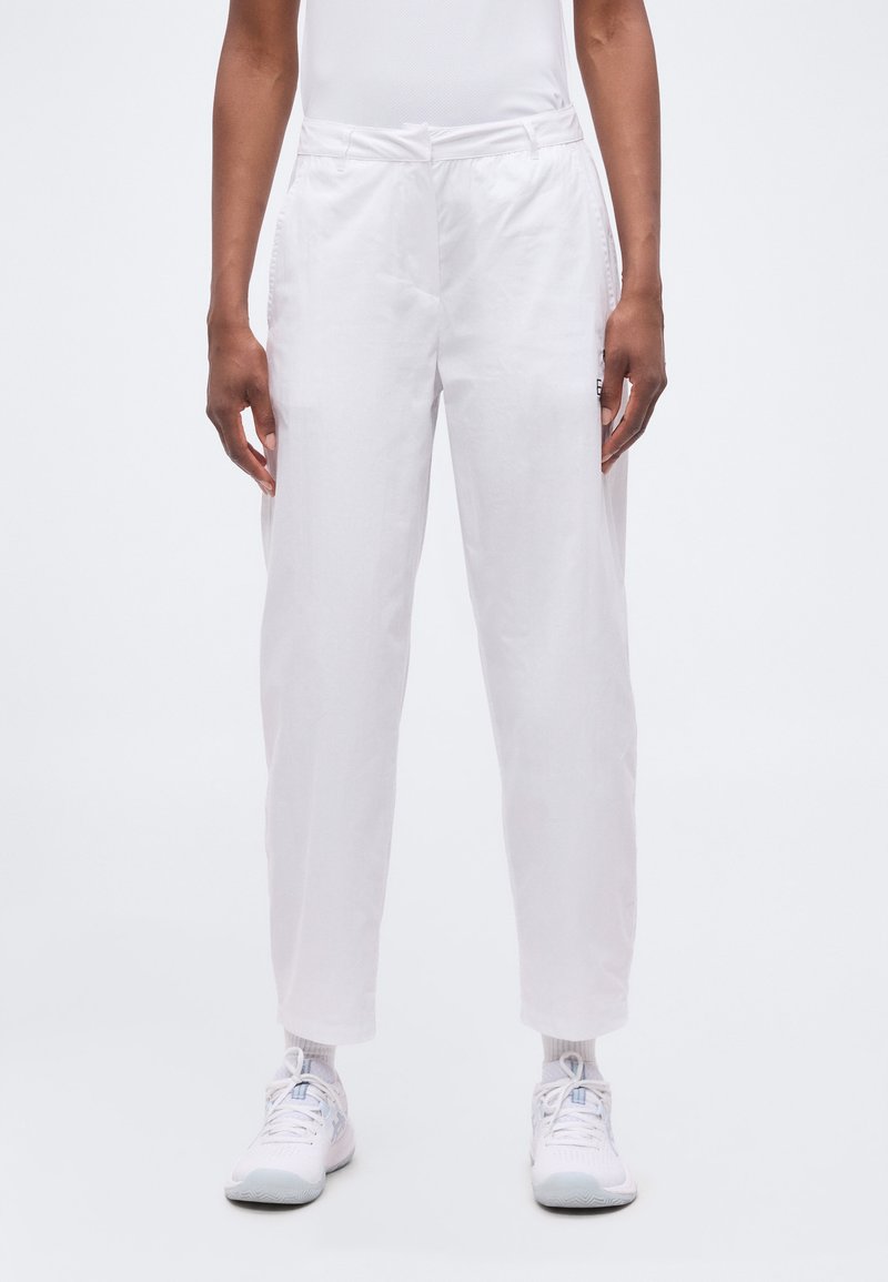 Άτομο που φορά φαρδιά λευκά παντελόνια, λευκές κάλτσες και λευκά αθλητικά παπούτσια, στέκεται μπροστά σε μια απλή φόντο.