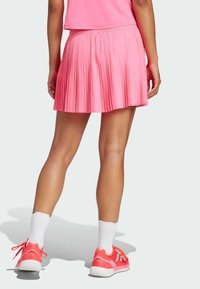 Falda plisada rosa con una cinturilla ancha, combinada con zapatillas rojas brillantes y calcetines blancos, vista desde atrás contra un fondo claro.