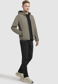 Helle olivfarbene Kapuzenjacke mit Reißverschluss vorne, elastischen Bündchen und Seitentaschen, kombiniert mit einem schwarzen Shirt und dunklen Jeans, getragen mit grauen Sneakern.