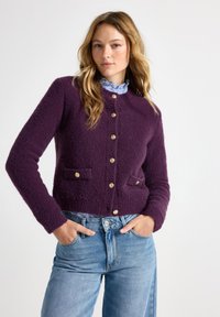 Purple textured cardigan with a round neckline, gold buttons, and two front pockets, paired with light denim jeans.