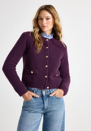 Purple textured cardigan with a round neckline, gold buttons, and two front pockets, paired with light denim jeans.