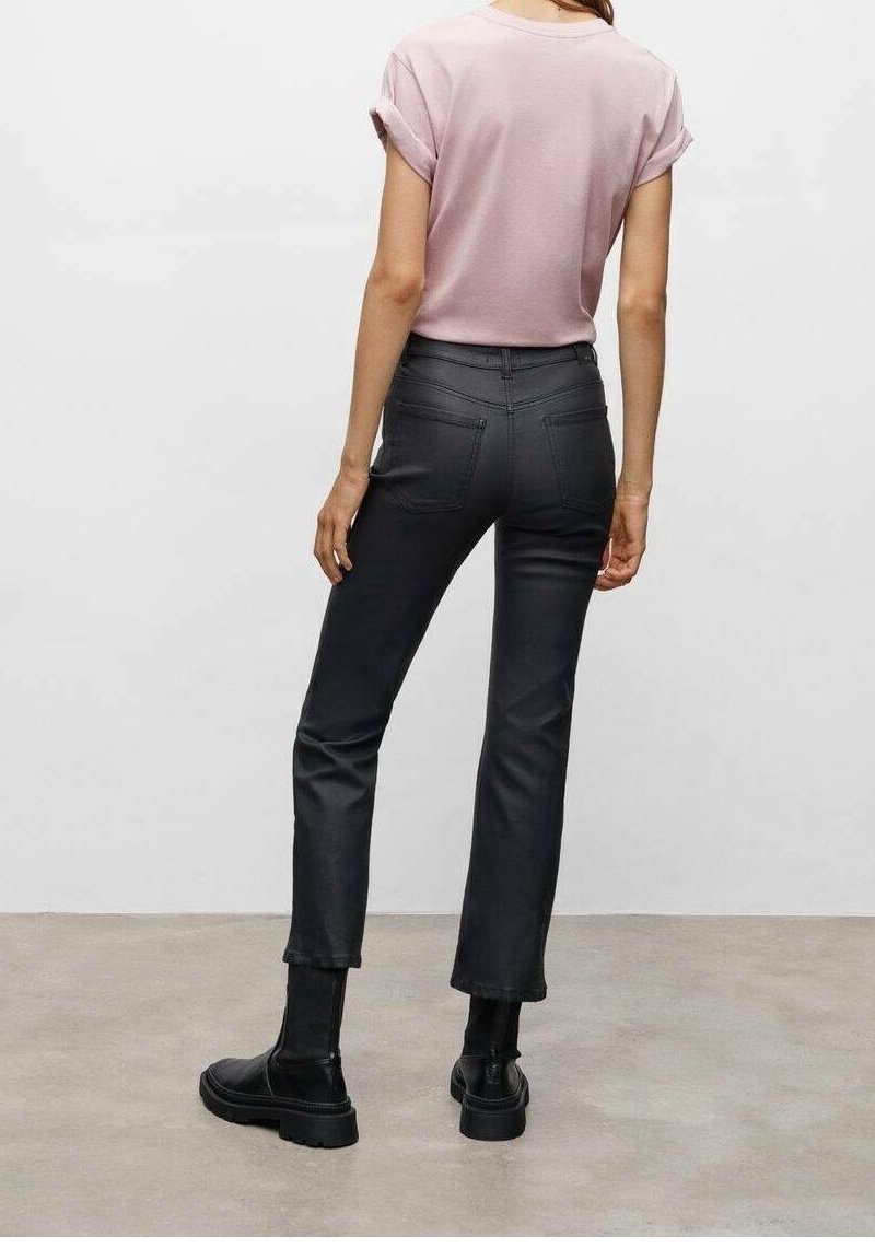 Femme portant une chemise rose clair à manches courtes, un pantalon court noir taille haute et des bottines noires, debout sur un sol gris.
