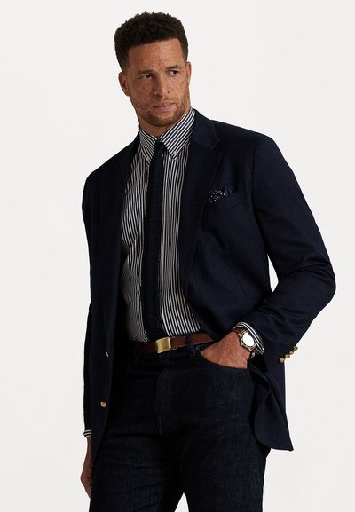 Ναυτικό σακάκι με χρυσά κουμπιά πάνω από πουκάμισο με κατακόρυφες ρίγες. Μαύρη γραβάτα και μαντηλάκι τσέπης, σκούρα τζιν παντελόνια. Ασημένιο ρολόι στον καρπό.