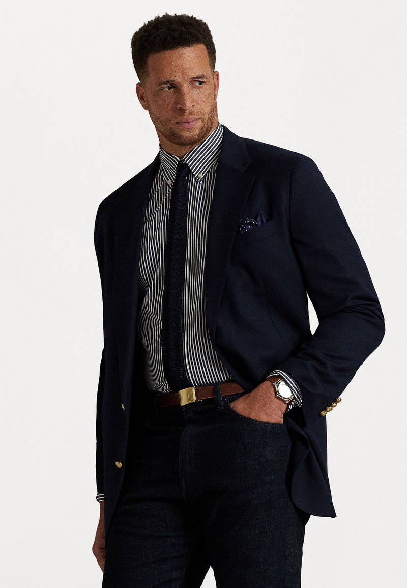 Blazer azul-marinho com botões dourados, camisa às riscas, gravata preta, cinto castanho, calças de ganga escuras e relógio prateado. Lenço de bolso acrescenta um toque de detalhe.