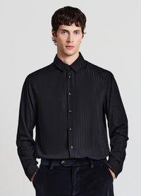 Camicia nera con bottoni davanti, a righe verticali sottili, texture liscia, colletto appuntito e bottoni neri. Indossata con pantaloni di velluto scuro.