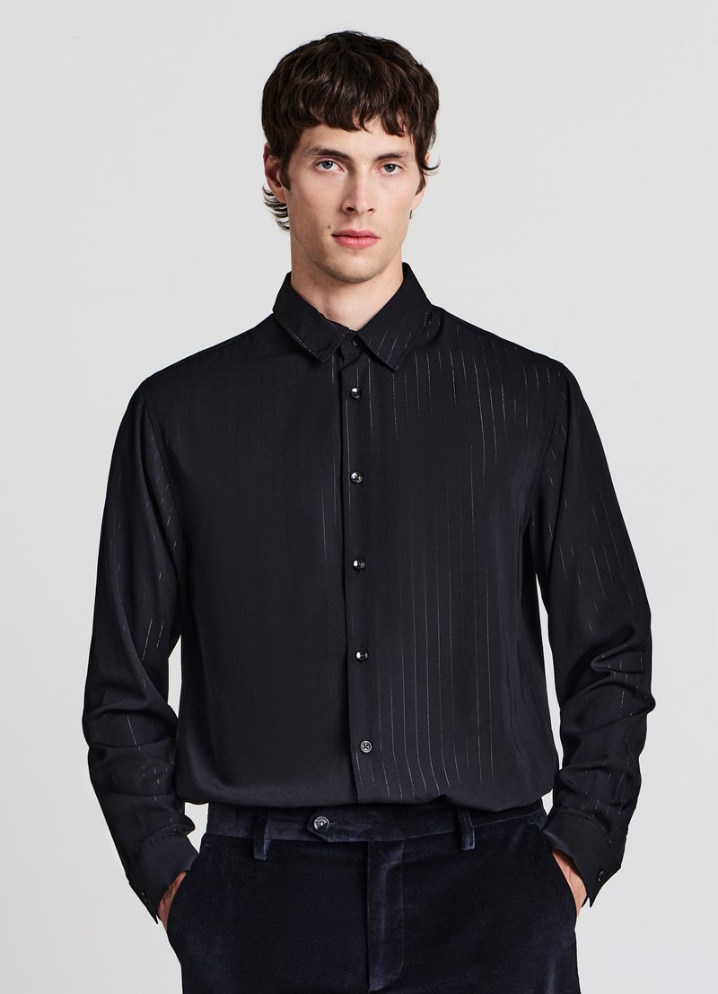 Camicia nera con bottoni davanti, a righe verticali sottili, texture liscia, colletto appuntito e bottoni neri. Indossata con pantaloni di velluto scuro.