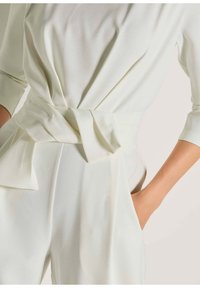 Lichtgroene jumpsuit met een zachte textuur, voorzien van een geknoopte taille, lange mouwen en zijzakken. Glad, vloeiend materiaal.
