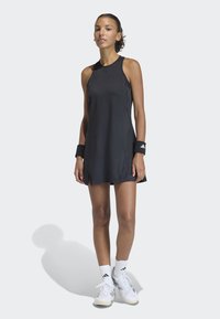 Athletische Frau in einem ärmellosen schwarzen Sportkleid, mit schwarzen Handgelenkbanden, weißen Socken und weißen Turnschuhen, die vor einem schlichten hellen Hintergrund steht.