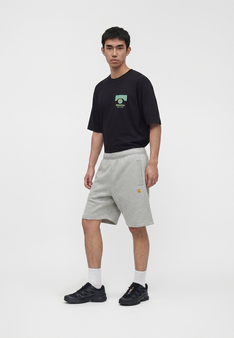 Junger Mann steht und trägt ein schwarzes T-Shirt mit Grafik, hellgraue knielange Shorts mit Logo, weiße Socken und schwarze Sneaker vor einfarbigem Hintergrund.