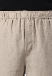 Beige linnen broek met een elastische tailleband, gedragen met een zwarte top, waarbij de textuur van de stof en de stiksels goed zichtbaar zijn.
