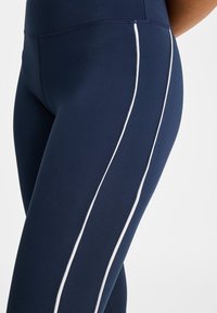 Marinblå leggings i ett stretchigt material med vita sidoremmar och hög midja. Texturen ser slät och matt ut.