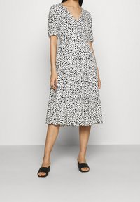 Krótkosleeve'owa sukienka z dekoltem w kształcie litery V w kolorze białym z czarnymi wzorami serc, warstwowym dołem i ściągniętą talią. Noszona z czarnymi sandałami na słupku.