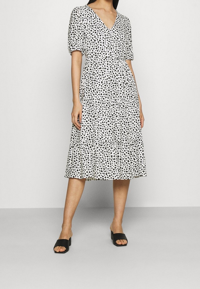 Krótkosleeve'owa sukienka z dekoltem w kształcie litery V w kolorze białym z czarnymi wzorami serc, warstwowym dołem i ściągniętą talią. Noszona z czarnymi sandałami na słupku.