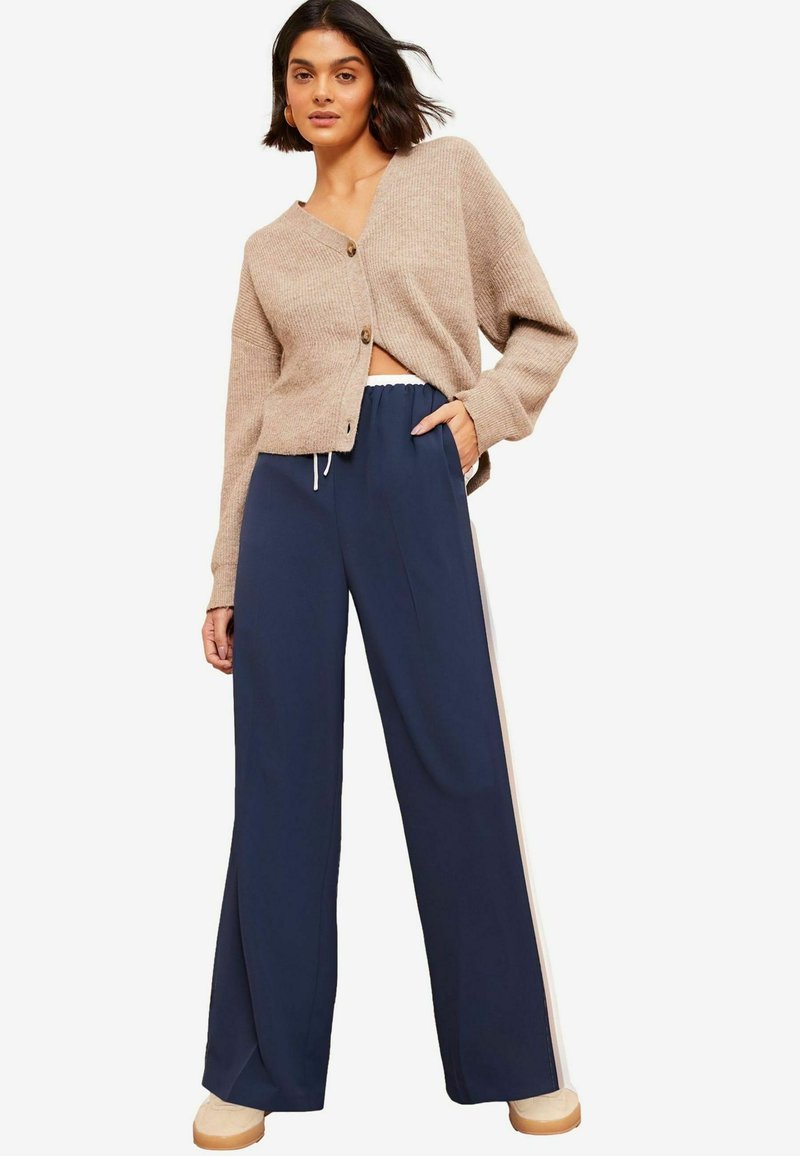 Cardigan en tricot beige avec une coupe courte, des manches oversized et une fermeture à boutons, associé à un pantalon large marine avec des rayures blanches sur les côtés.