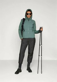 Fleecejakke i grønn med hette, sorte isolerte bukser og turstaver. Teksturert stoff og funksjonelle glidelåser er synlige.