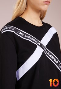 Mujer vestida con una sudadera negra con franjas blancas entrecruzadas y el texto "KARL LAGERFELD" en el pecho y el hombro.
