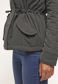 Gros plan d'une personne portant une veste vert foncé à cordon de serrage à la taille et poche à rabat, associée à un jean bleu.