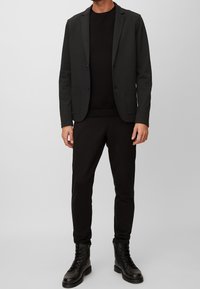 Man som bär en svart blazer, svart tröja, svarta slim-fit byxor och svarta snörkängor, stående mot en ljusgrå bakgrund.