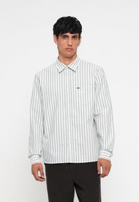 Camisa de botones con rayas verticales en gris claro y blanco, mangas largas y un pequeño logo bordado en el bolsillo del pecho.