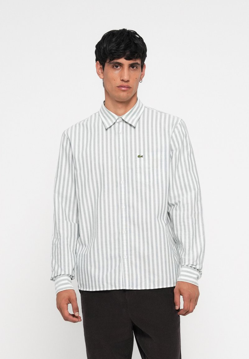 Camisa de botones con rayas verticales en gris claro y blanco, mangas largas y un pequeño logo bordado en el bolsillo del pecho.
