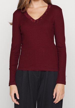 Vrouw draagt een bordeauxrode geribbelde top met lange mouwen en een kanten randje aan de V-hals, gecombineerd met losse zwarte broek.