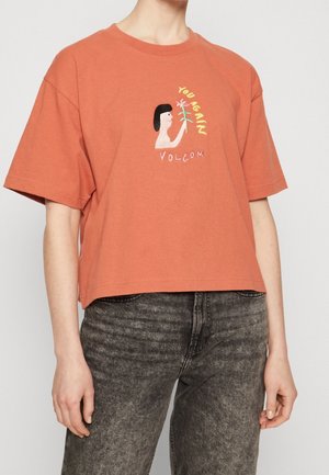 Osoba nosząca pomarańczowy krótki t-shirt z ilustracją kobiety trzymającej kwiat oraz napisami "YOU GAIN" i "VOLCOM" na przodzie.