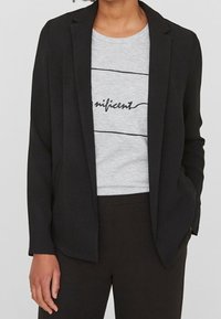 Czarna marynarka o dopasowanym kroju, z klapami, przednimi kieszeniami, noszona na szarym t-shircie z czarnym tekstem pisanym kursywą i poziomymi liniami.