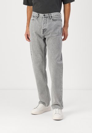 Personne portant un jean droit gris clair avec une fermeture éclair et un bouton, associé à des baskets blanches et un haut noir.