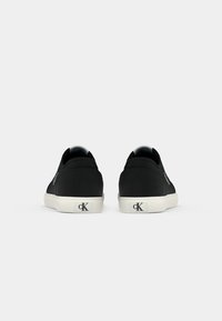 Para czarnych niskich sneakerów z białymi podeszwami i logo "cK" na pięcie, pokazanych z tyłu na jednolitym jasnym tle.
