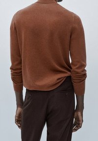 Uomo che indossa un maglione color ruggine con le maniche arrotolate e pantaloni marroni scuri, visto di spalle su uno sfondo neutro.