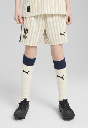 Beige sportshorts med vertikala ränder, med marinblå detaljer och en logotyp. Svarta fotbollsskor med strukturerad yta; knähöga krämfärgade strumpor med marinblå band.