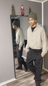 Felpa con cappuccio a quadri beige con rifiniture nere, abbinata a pantaloni neri larghi. Berretto lavorato a maglia verde. Lo specchio a figura intera riflette il modello in una stanza neutra.