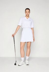 Valge golfipolo ja seelik, mõlemad hingavast kangast, kombineeritud valgete spordijalatsite ja sokkidega. Hoidmas golfikeppi.