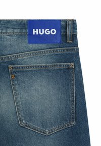 Denim jeans featuring a blue rectangular label with "HUGO" in white, visible stitching, and a back pocket with a decorative stitch detail.