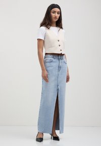 Gilet sans manches beige avec des détails de boutons, associé à un t-shirt blanc et une jupe maxi en denim bleu clair taille haute avec une fente à l'avant. Escarpins noirs.