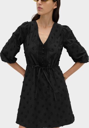 Donna che indossa un mini abito nero testurizzato con maniche a sbuffo, davanti con bottoni, cintura in vita e occhiali da sole con lenti gialle, mani sui fianchi.