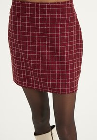 Mini falda a cuadros burdeos con patrón de rejilla blanca; tejido texturizado; diseño ajustado; dobladillo por encima de los muslos; combinada con medias negras y botas claras.