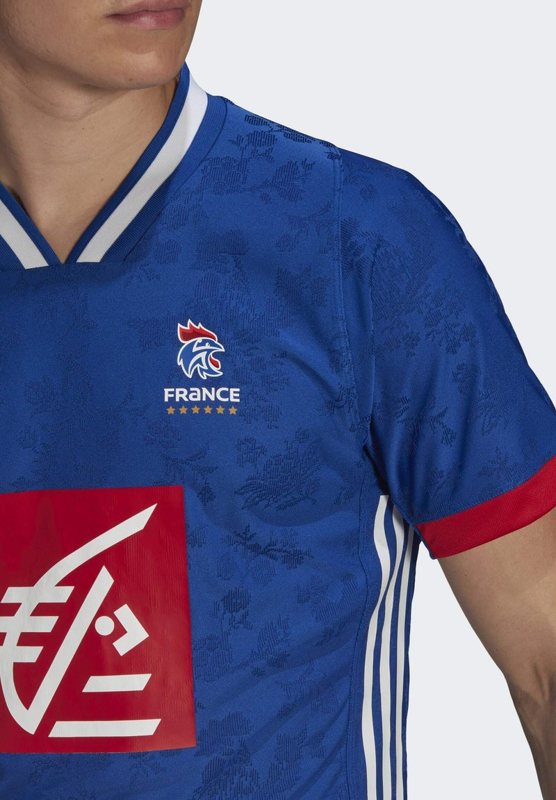 Blauw sportshirt met een subtiel bloemmotief, rode accenten op de mouwen, een witte gestreepte kraag en een prominent logo met tekst.