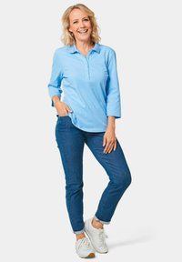 Helleblaue Langarm-Poloshirt mit drei Knöpfen, kombiniert mit dunkelblauen Jeans und weißen Sneakers mit strukturierten Akzenten.