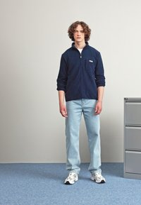 Giacca a zip in pile navy con tasca frontale, jeans azzurri chiari e scarpe da ginnastica bianche. Texture liscia, vestibilità casual, branding minimo.