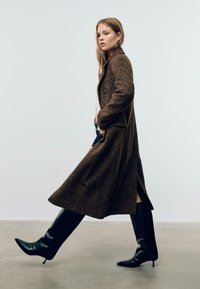 Manteau long en chevrons brun avec un bouton unique et des fentes latérales, porté sur un haut léger, associé à des bottes noires à talons montantes.