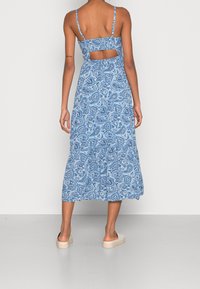 Modrá maxi šaty s paisley vzorem a výstřihem na zádech, tenkými ramínky a odpovídajícími plochými sandály na hladké, světlé podlaze.