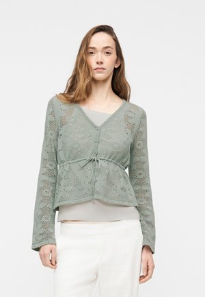 Joven mujer con un cárdigan de encaje tejido de color verde claro con botones y lazo en la cintura, sobre una camiseta clara, combinado con pantalones blancos.