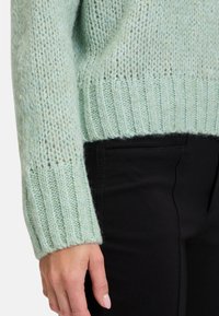 Ljusgrön stickad tröja med ribbade manschetter och fåll, med en mjuk textur och lös passform. Bärs över svarta byxor med fickor.