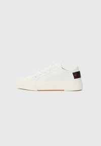 Sneaker de lona branca com biqueira redonda, sola de borracha plana e uma aba preta no calcanhar com um emblema de coração vermelho. Ilhós para os atacadores na parte da frente.